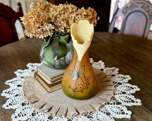 A vase made from a dried gourd. It is a brown to green ombre stain, with a iridescent butterfly wood burned on the front.  